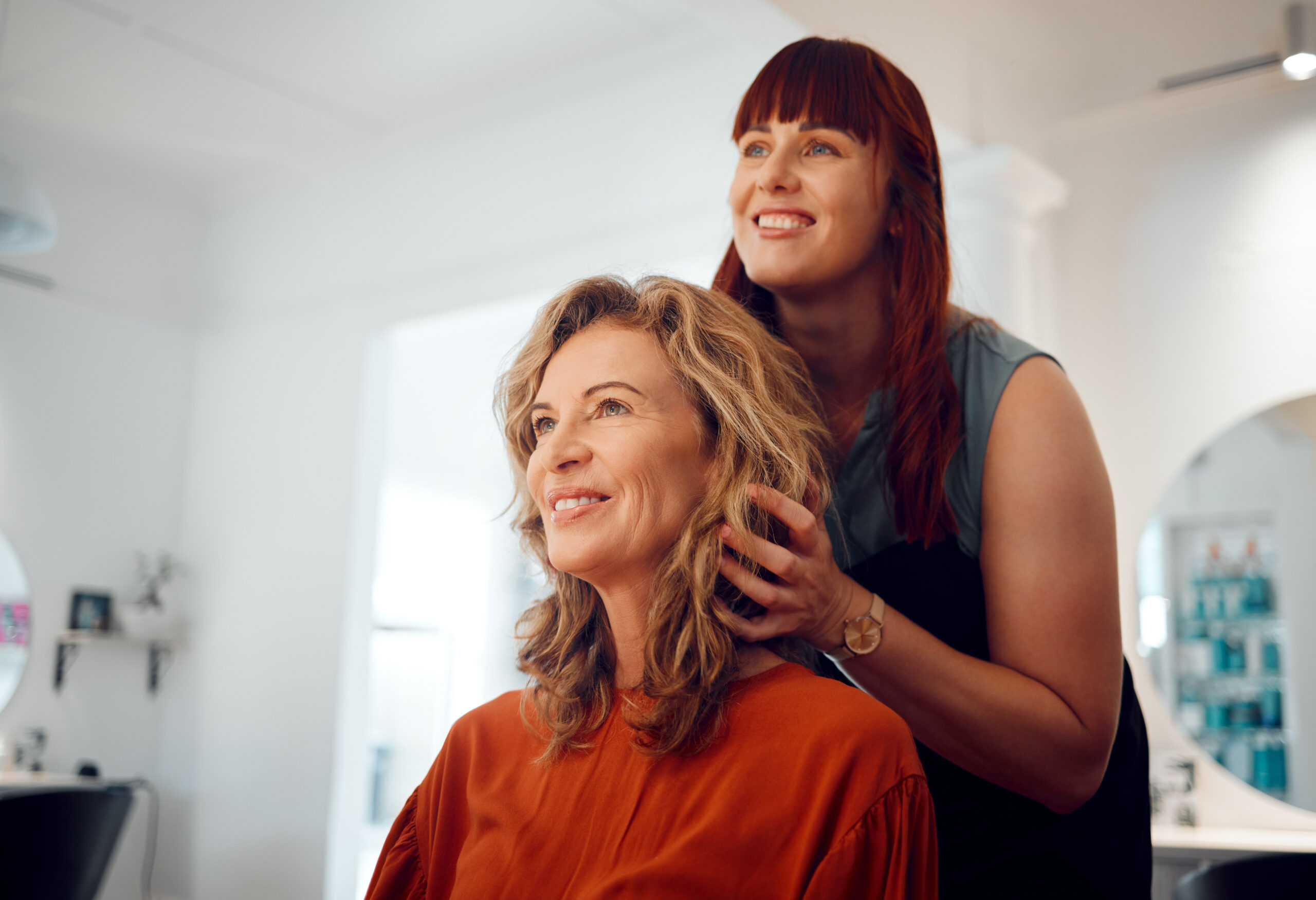 Eine junge, rothaarige Friseurin stylt ihre blonde, etwas ältere Kundin. Beide lächeln freudig in die Kamera und freuen sich über die neue Frisur